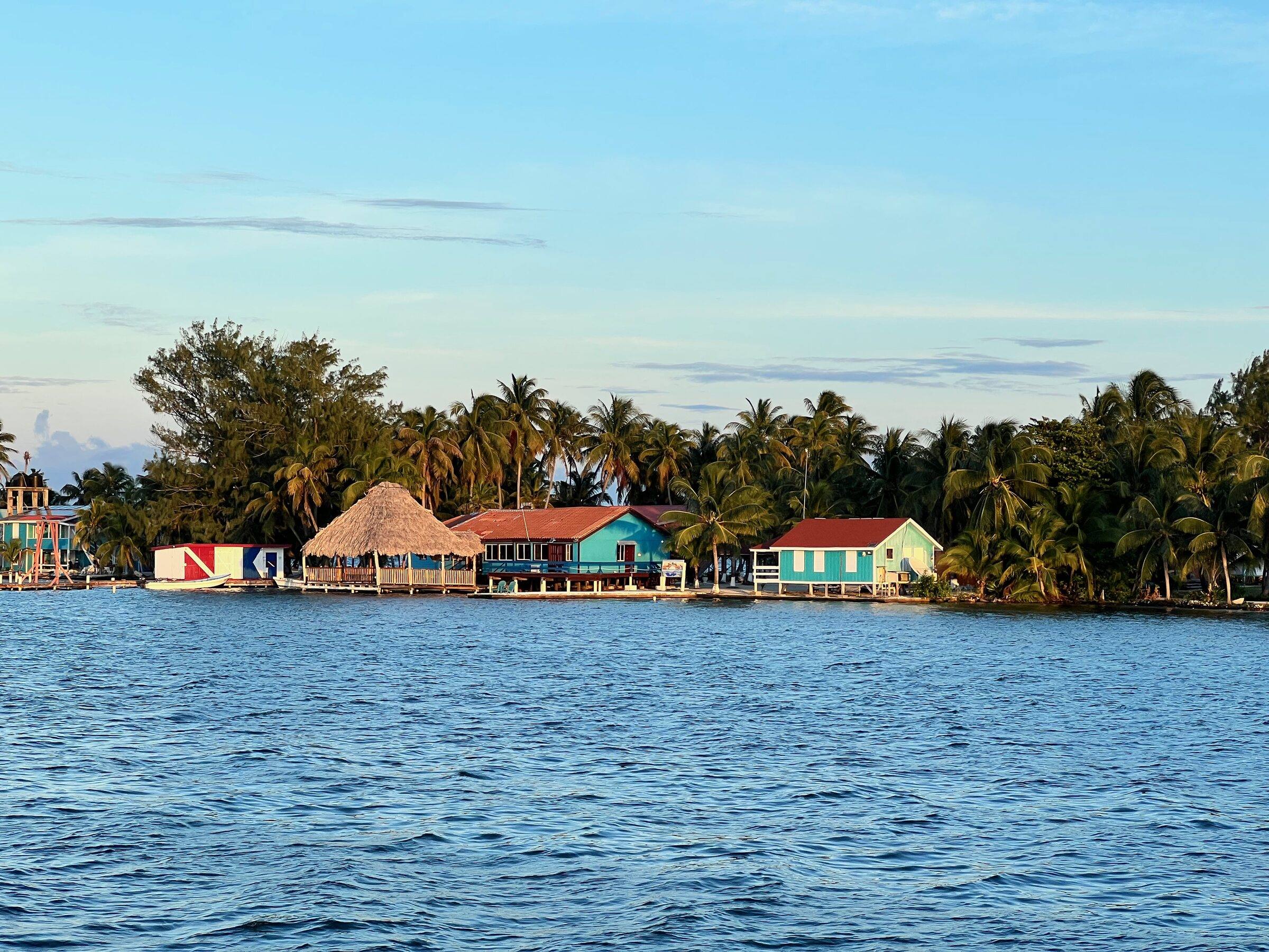 Colourful wooden buildings and a thatched hut among palms on a small caye, seen from the water
