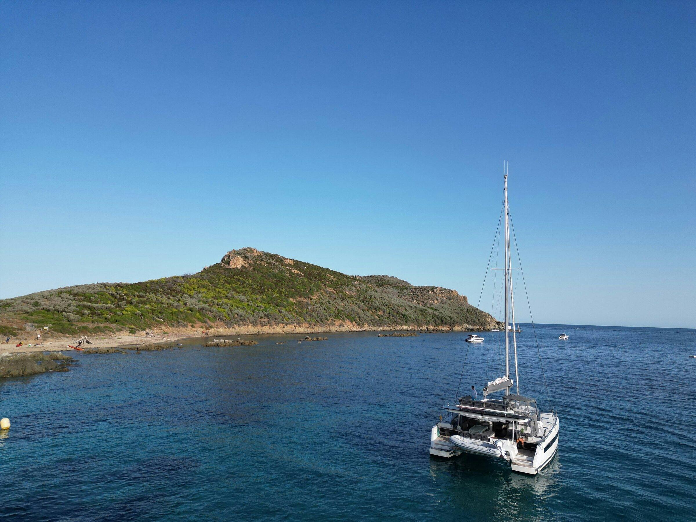 Catamaran at anchor off a green, rocky headland on calm blue sea