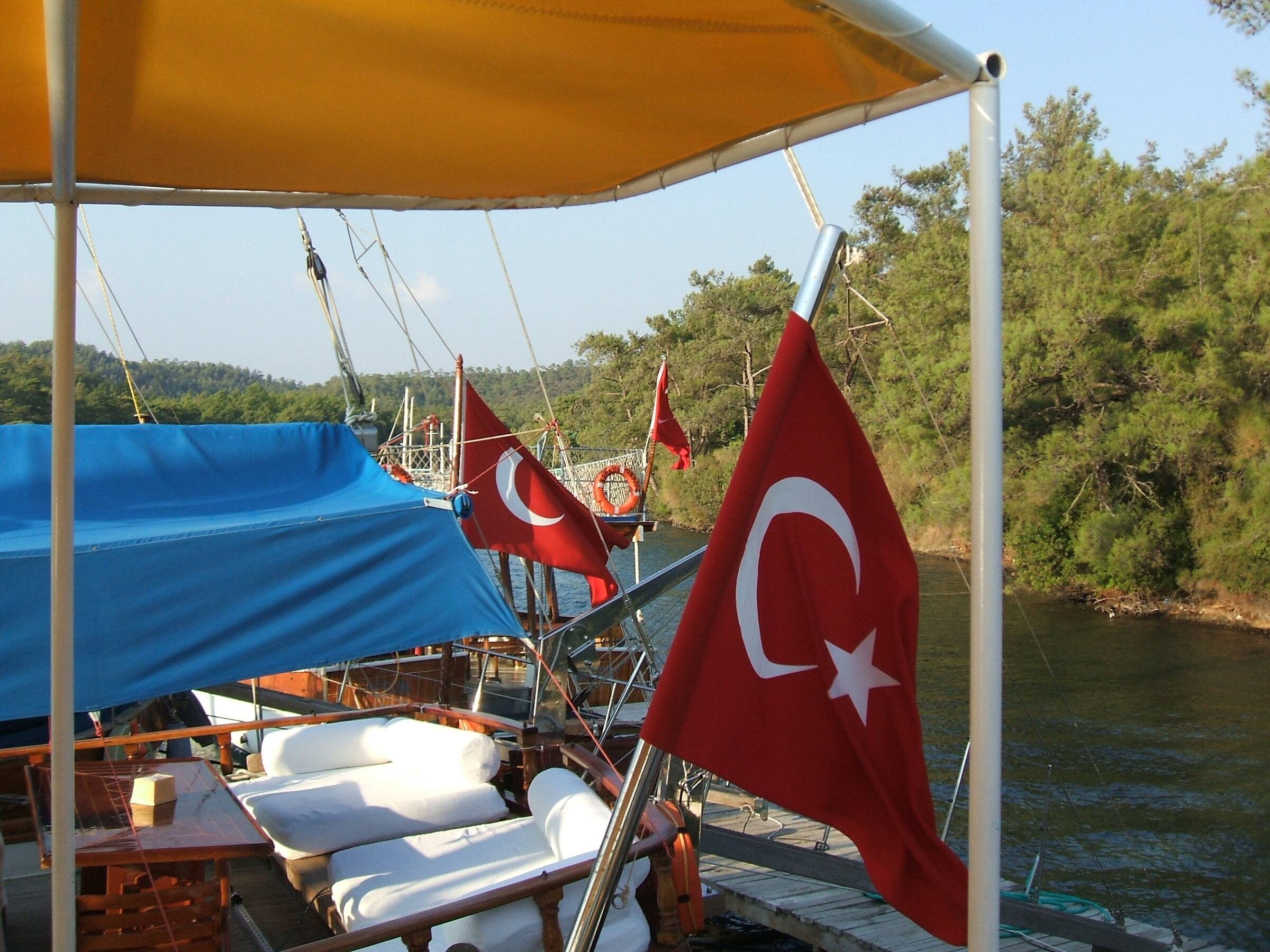 Cushioned lounge area on the deck of a gulet under a blue awning, with Turkish flags flying