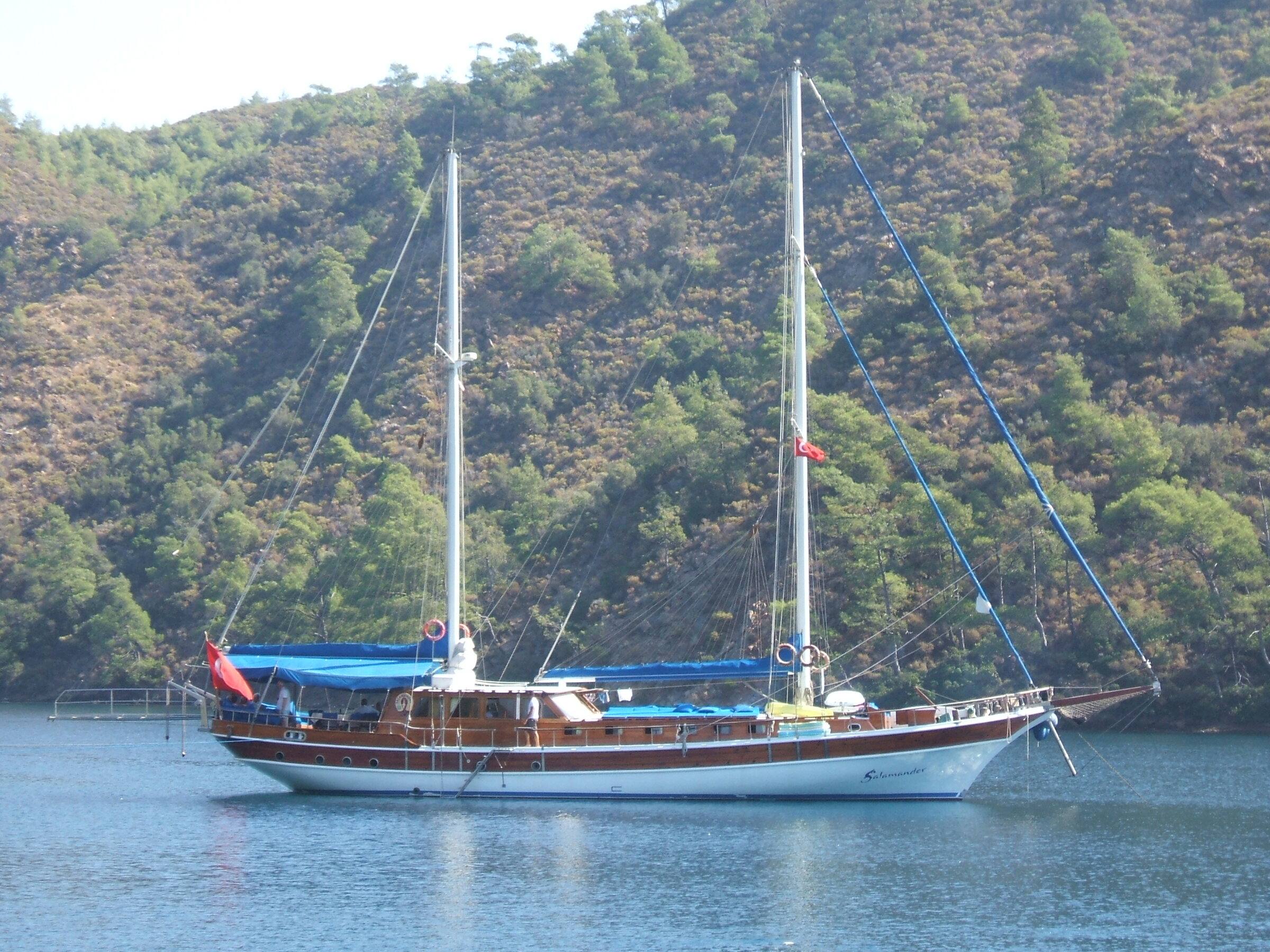 Gulet, a traditional wooden two-masted boat with a blue awning, at anchor in a calm wooded bay
