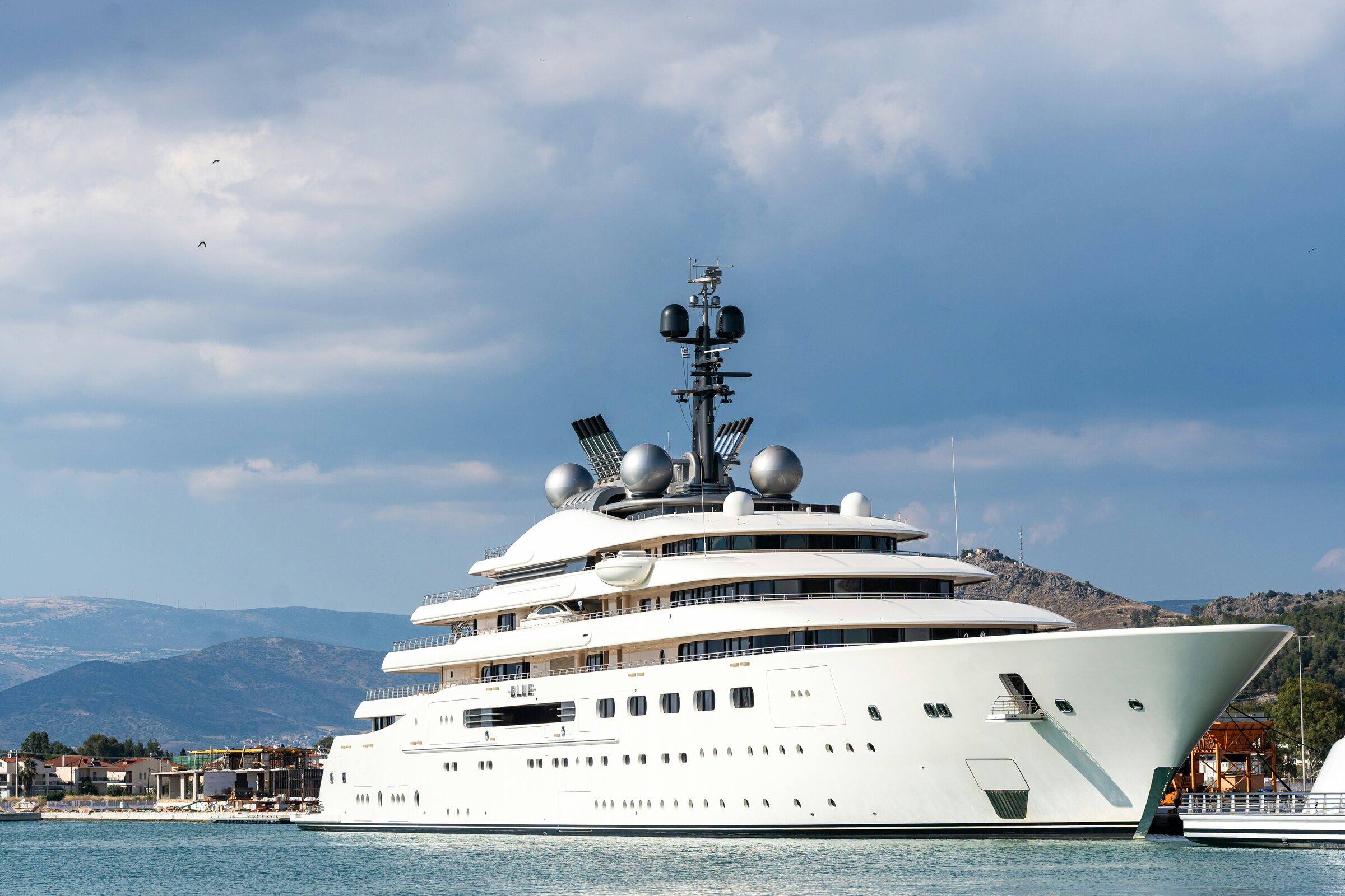 Large white motor yacht moored in a bay under a cloudy sky, with hills behind