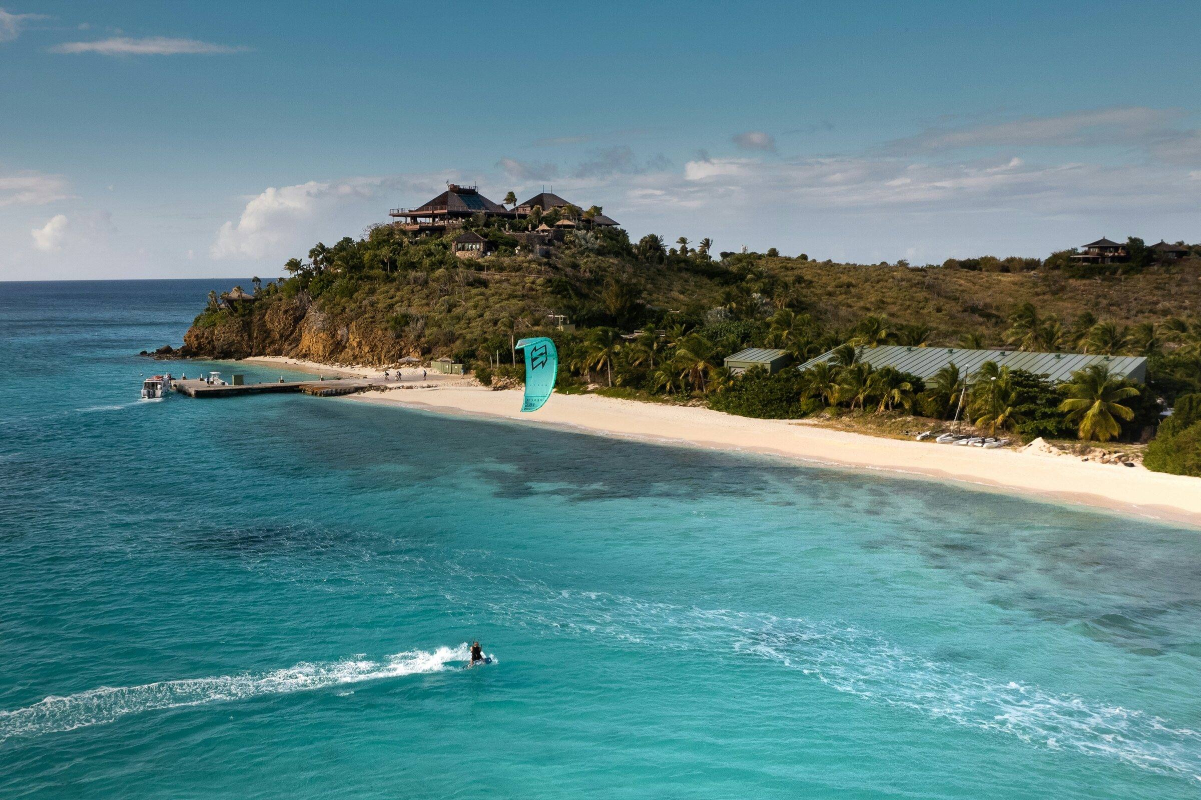Aerial view of a sandy beach and palms on a green headland beside turquoise water, with a kiteboarder on the water