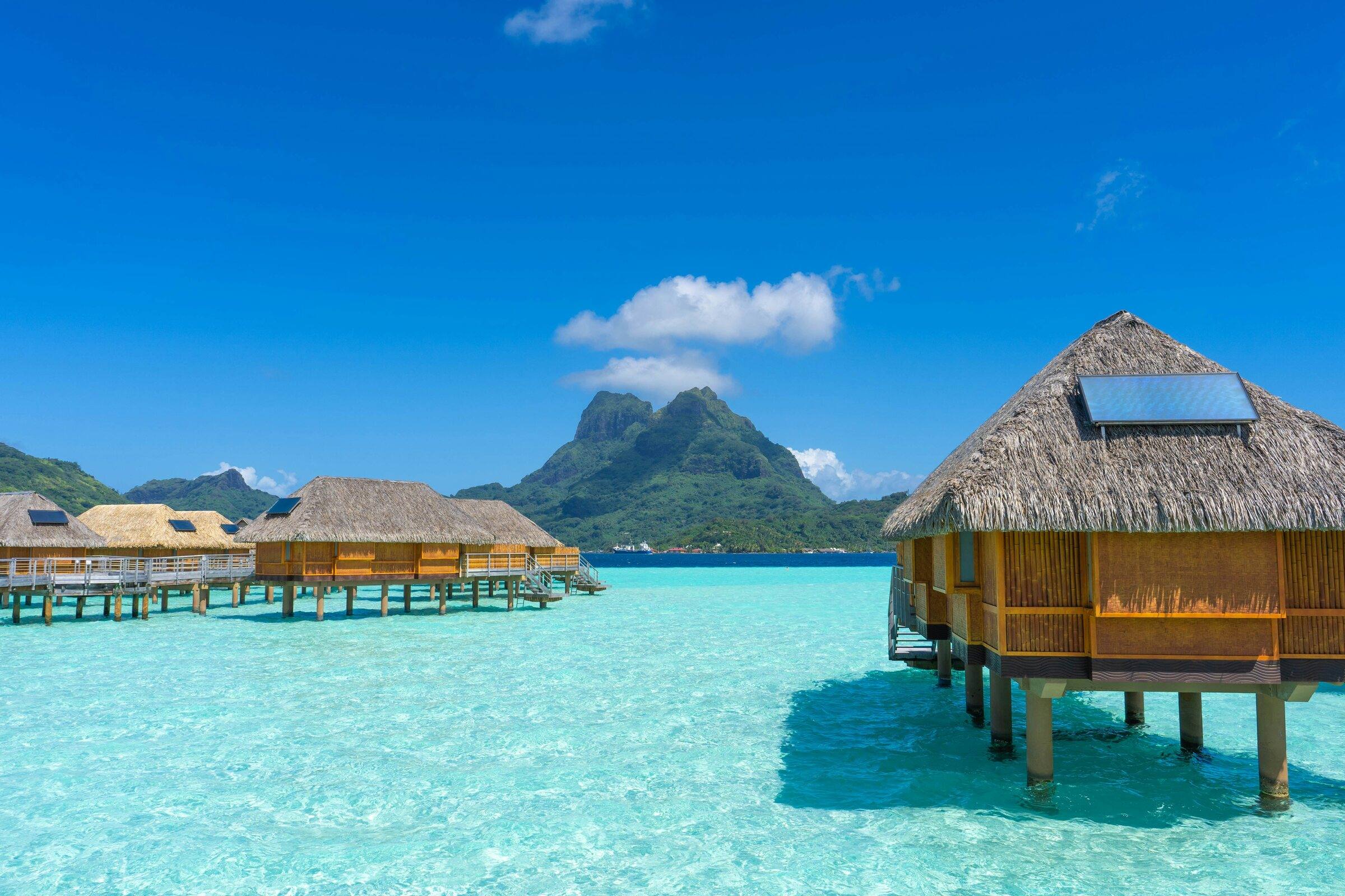 Thatched overwater bungalows on a clear turquoise lagoon with a green volcanic peak behind
