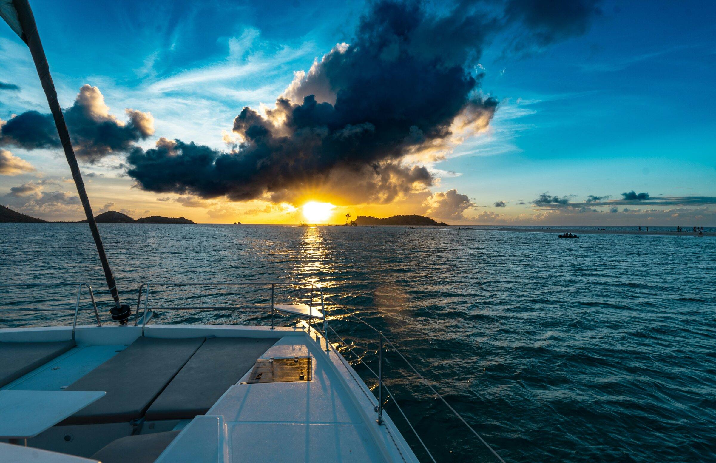 Sunset over calm sea seen from the bow of a catamaran, with low islands on the horizon