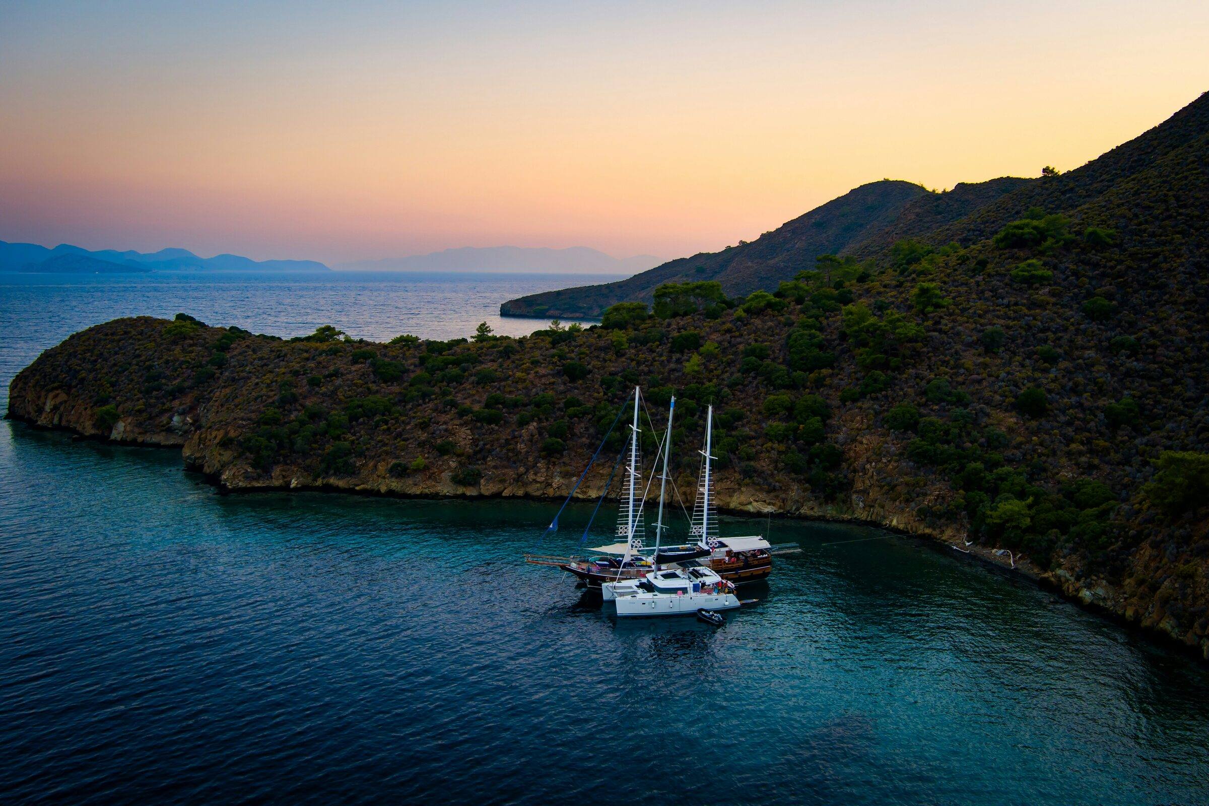 Two traditional wooden two-masted boats and a white catamaran at anchor in a wooded bay at dusk